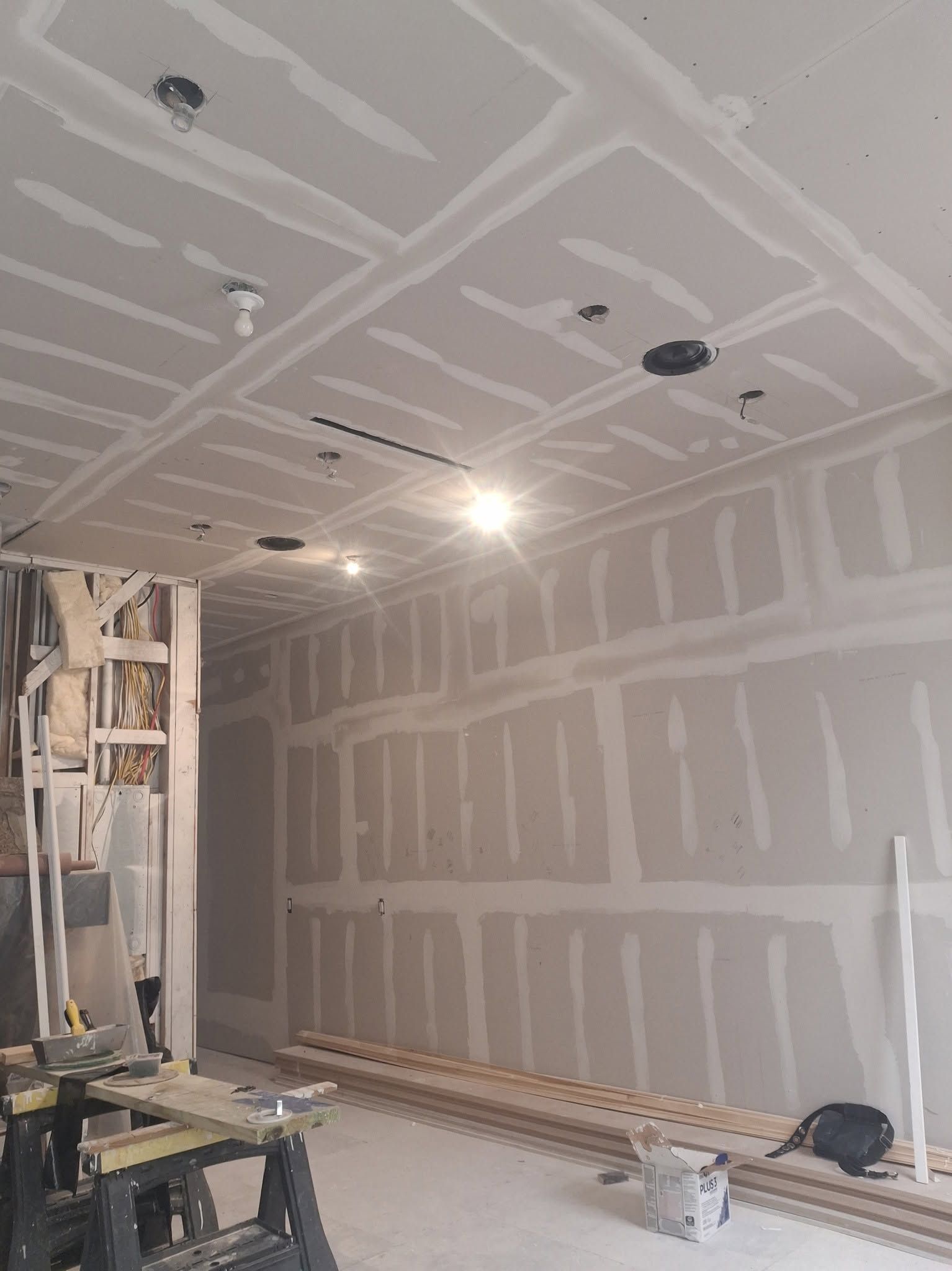 A room with sheetrock walls and ceiling, the seams taped and mudded, tools on the floor, before paint.